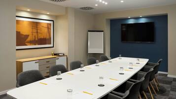 Uma sala de conferências com uma mesa comprida, cadeiras, um quadro branco e uma televisão na parede.