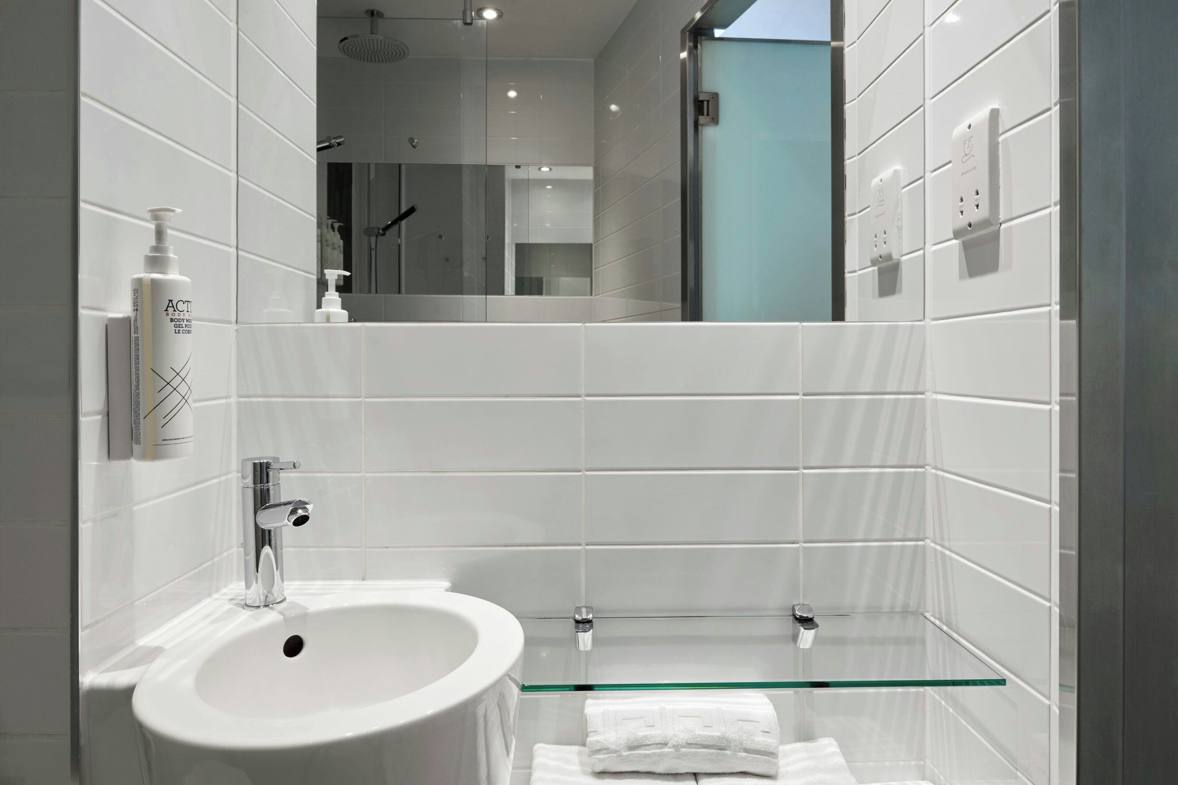 Une salle de bain moderne avec un lavabo blanc, une étagère en verre et un miroir qui reflète l’espace de la douche.