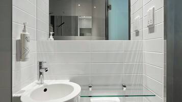 Une salle de bain moderne avec un lavabo blanc, une étagère en verre et un miroir qui reflète l’espace de la douche.