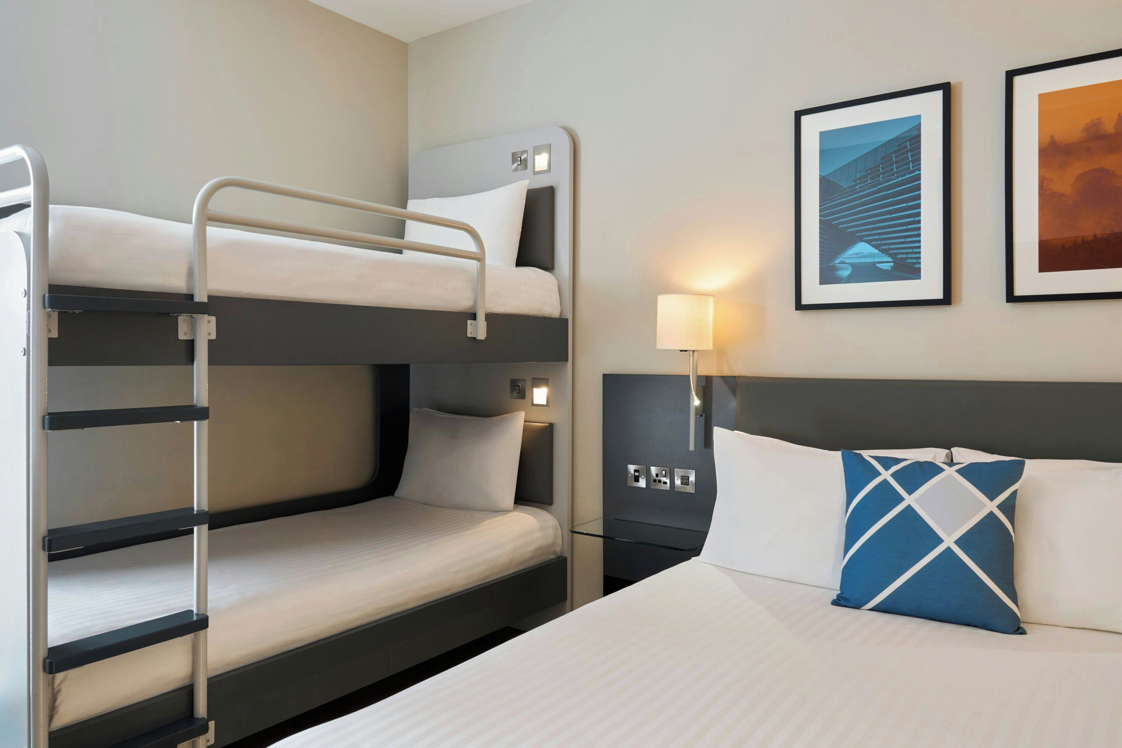 Een hotelkamer met stapelbedden, een nachtkastje met een lamp en ingelijste foto’s aan de muur.