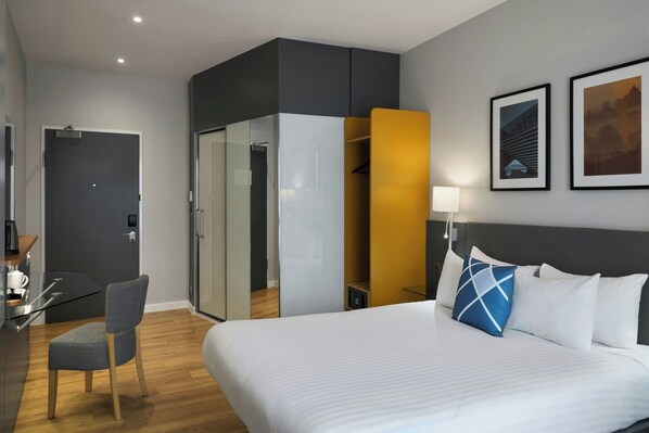 Una habitación de hotel moderna con una cama grande, un escritorio con silla, un armario con puertas de vidrio y cuadros en la pared.