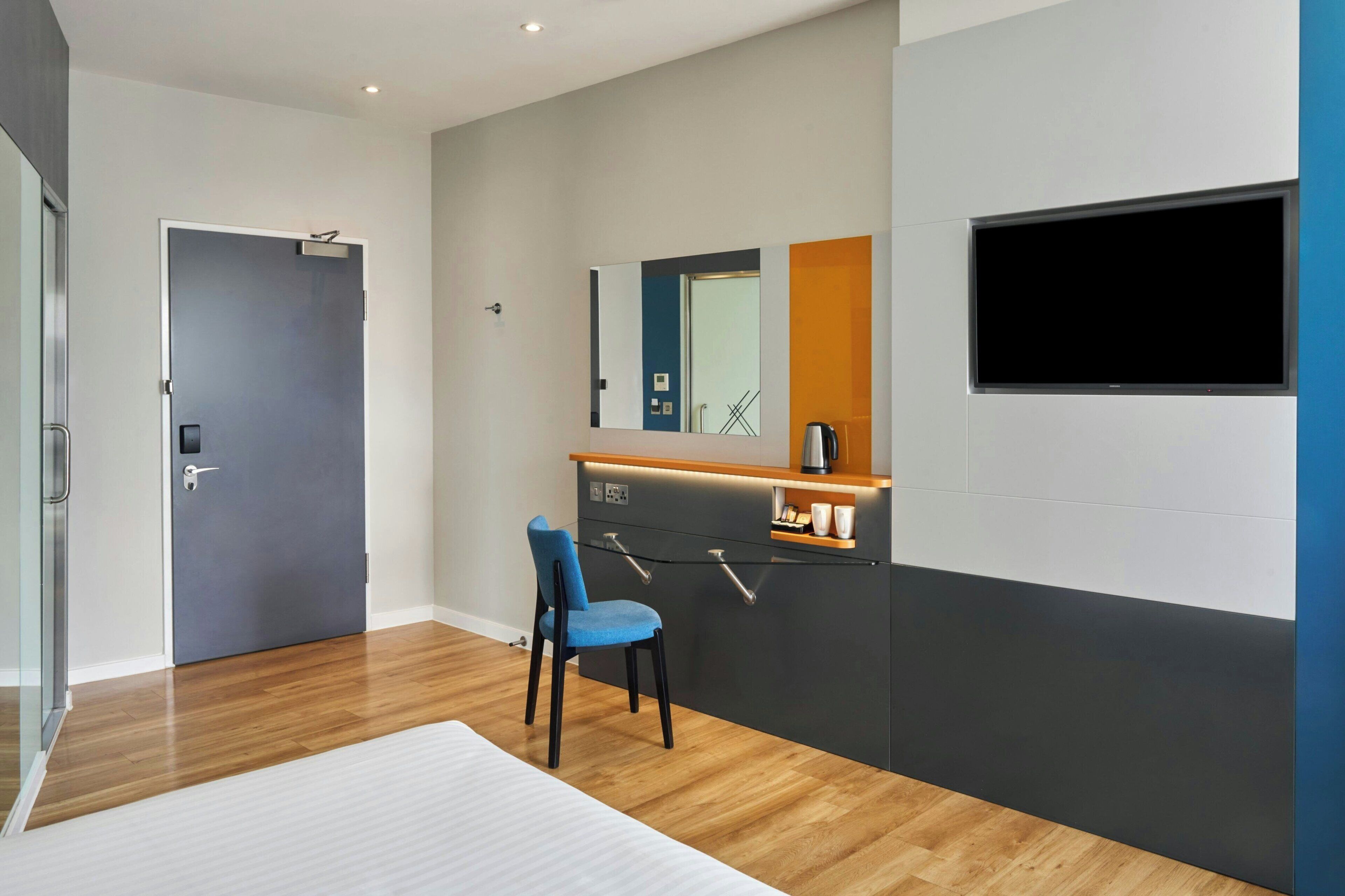 Una habitación de hotel moderna con suelo de madera, una silla azul, un armario oscuro y un televisor de pantalla plana montado en la pared, junto a una puerta con manija.