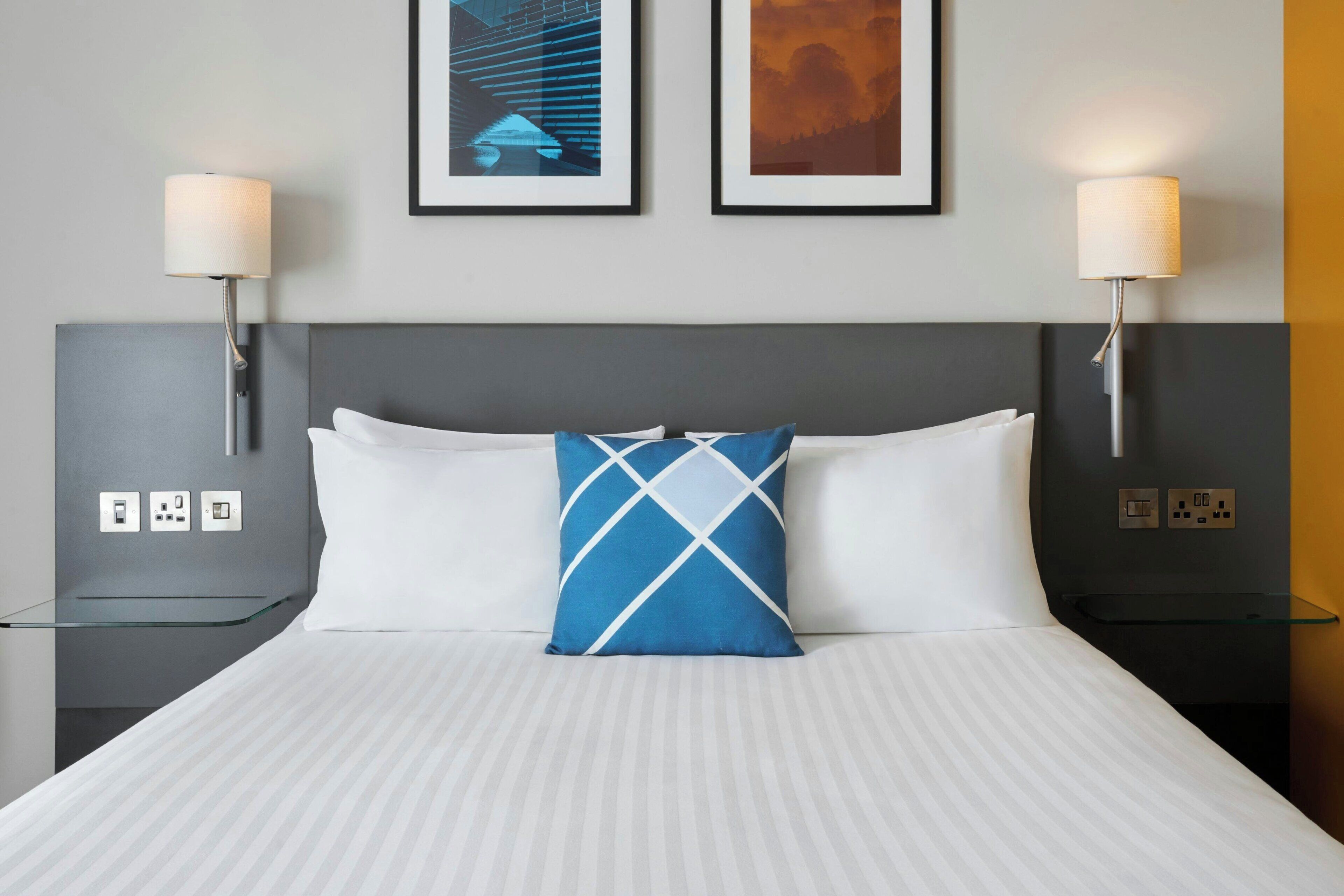 A hotel room with a bed, two bedside lamps, a framed picture on the wall, and a glass table.