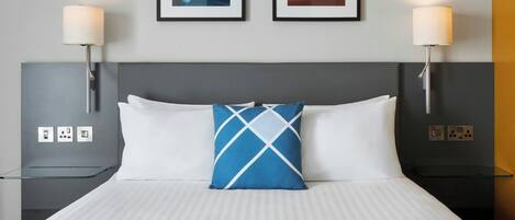 A hotel room with a bed, two bedside lamps, a framed picture on the wall, and a glass table.