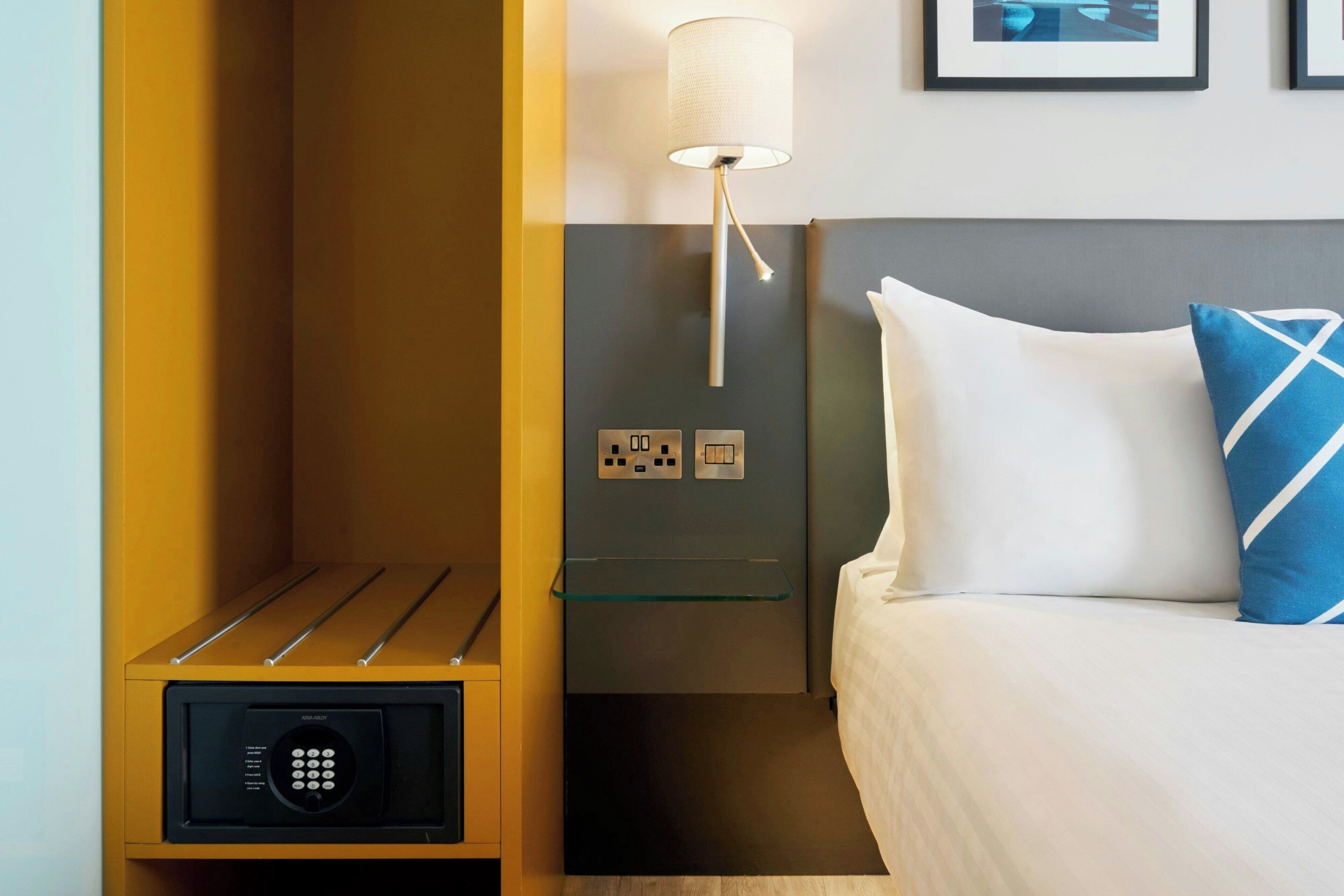 Een hotelkamer met een bed, een bedlampje, een glazen plank, een kluis en stopcontacten.