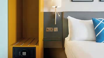 Een hotelkamer met een bed, een bedlampje, een glazen plank, een kluis en stopcontacten.