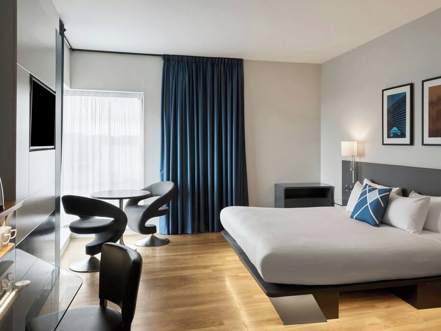 A modern hotel room with a bed, a desk, a chair, a coffee maker, and a lamp.