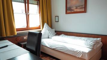 Habitación de hotel con cama, escritorio, silla, ventana con cortinas amarillas y un cuadro en la pared.