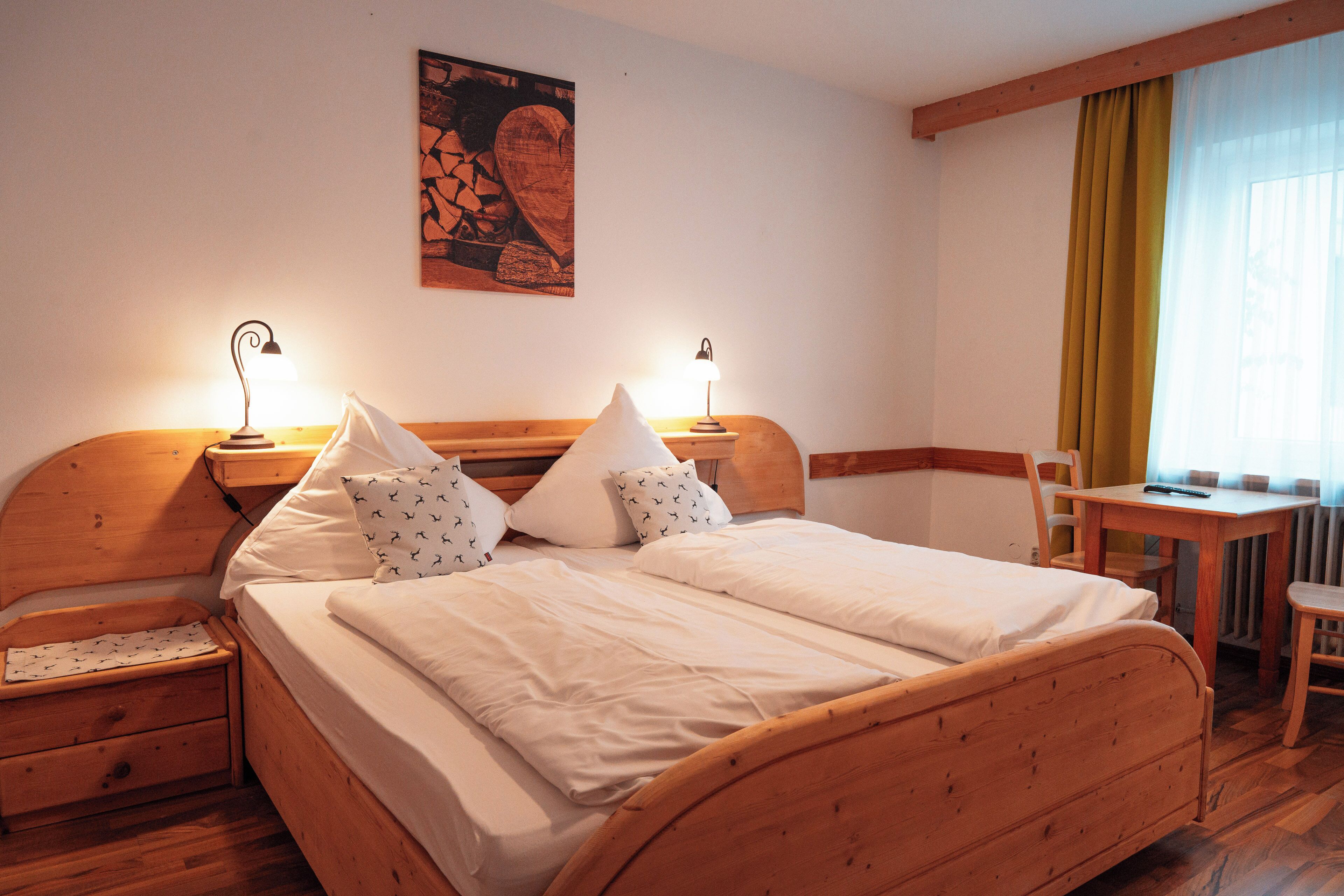 A wooden bed with white linens and two bedside lamps, a wooden nightstand with a drawer, a wooden chair, and a table with a chair.
