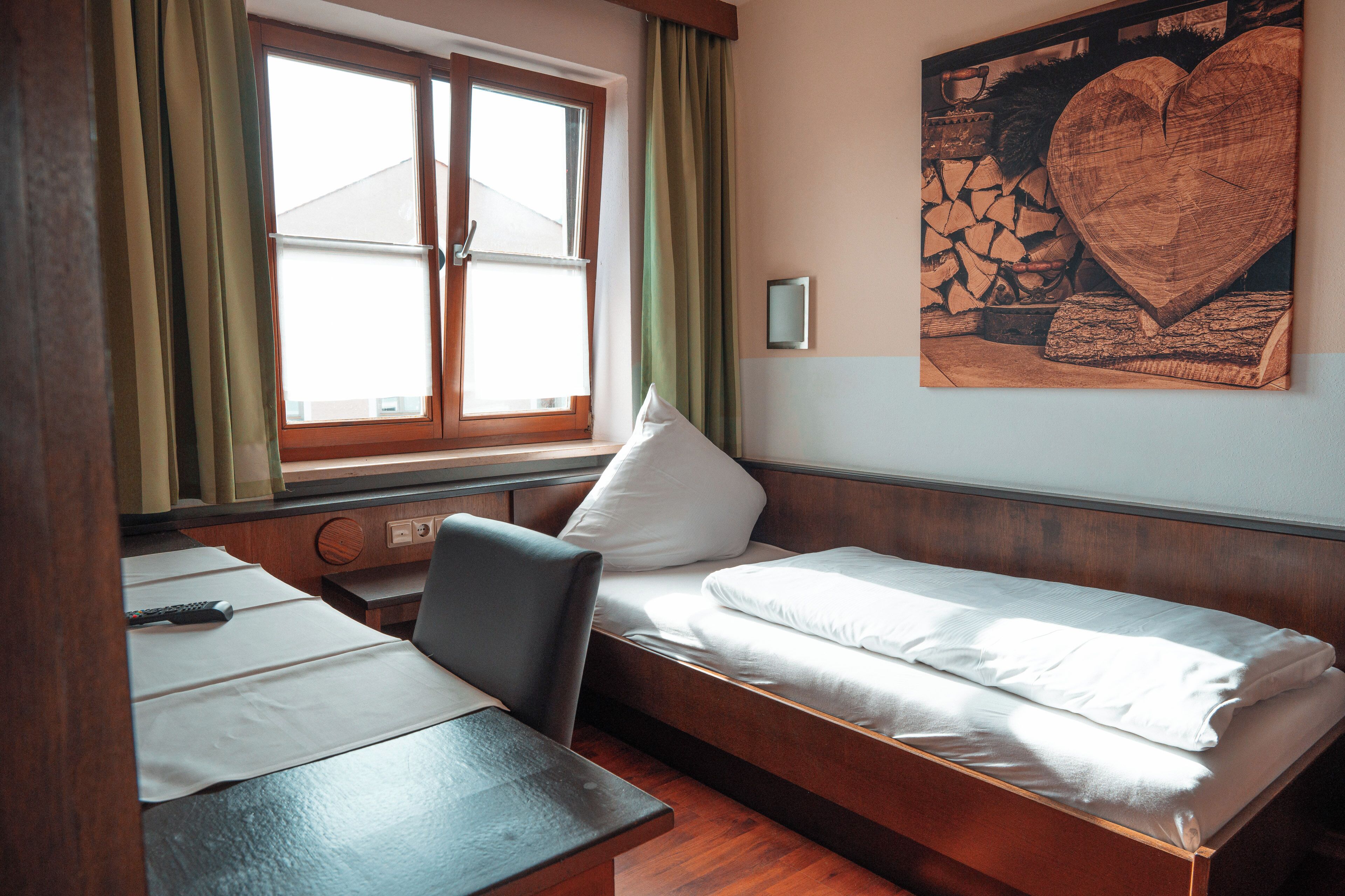 Ein Einzelbett mit Holz-Kopfstück, ein Nachttisch, ein Stuhl und ein Fenster mit Vorhängen.