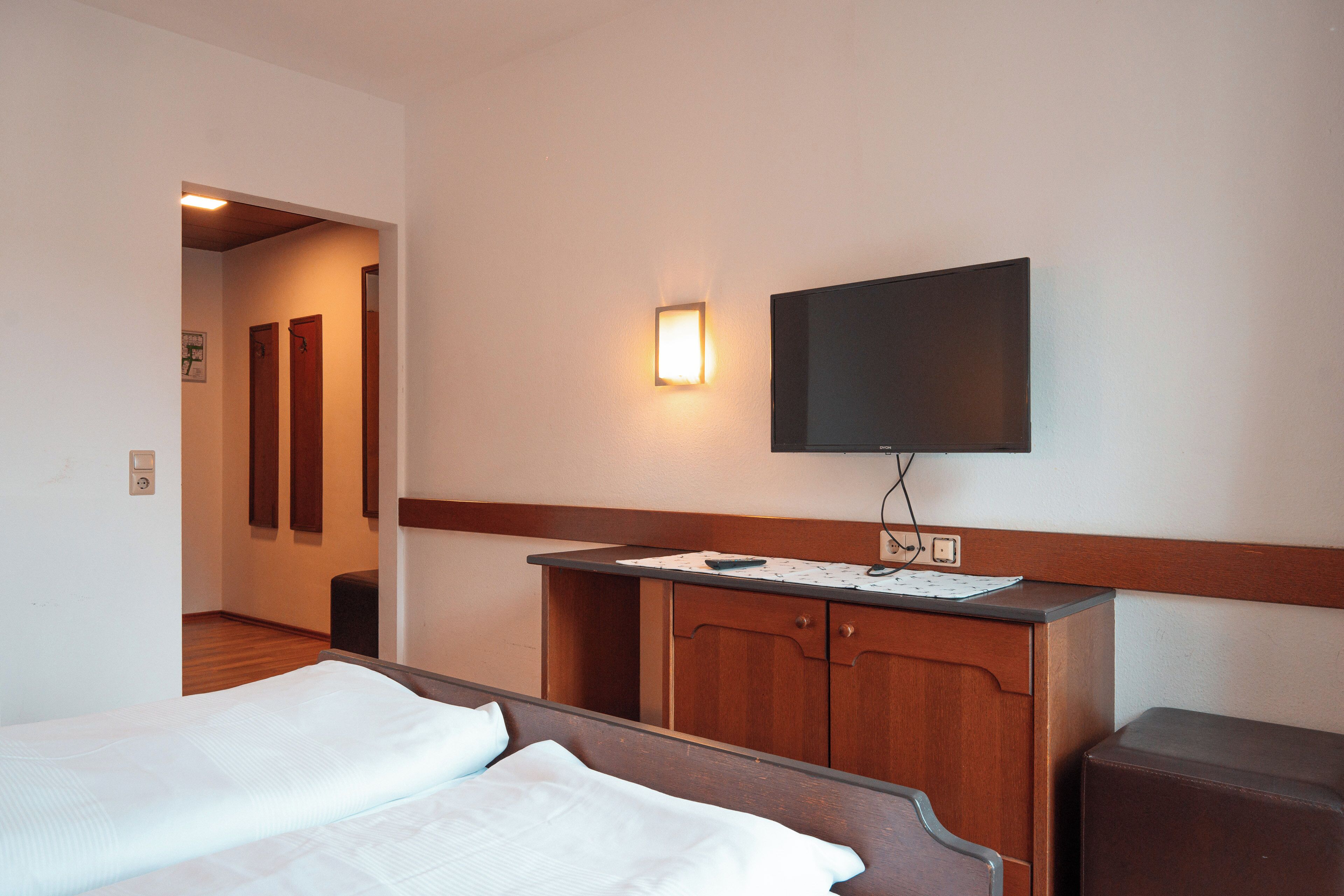 Ein Hotelzimmer mit einem Bett, einer Holzkommode, einem flachbildfernseher an der Wand und einem kleinen Schreibtisch mit Lampe.