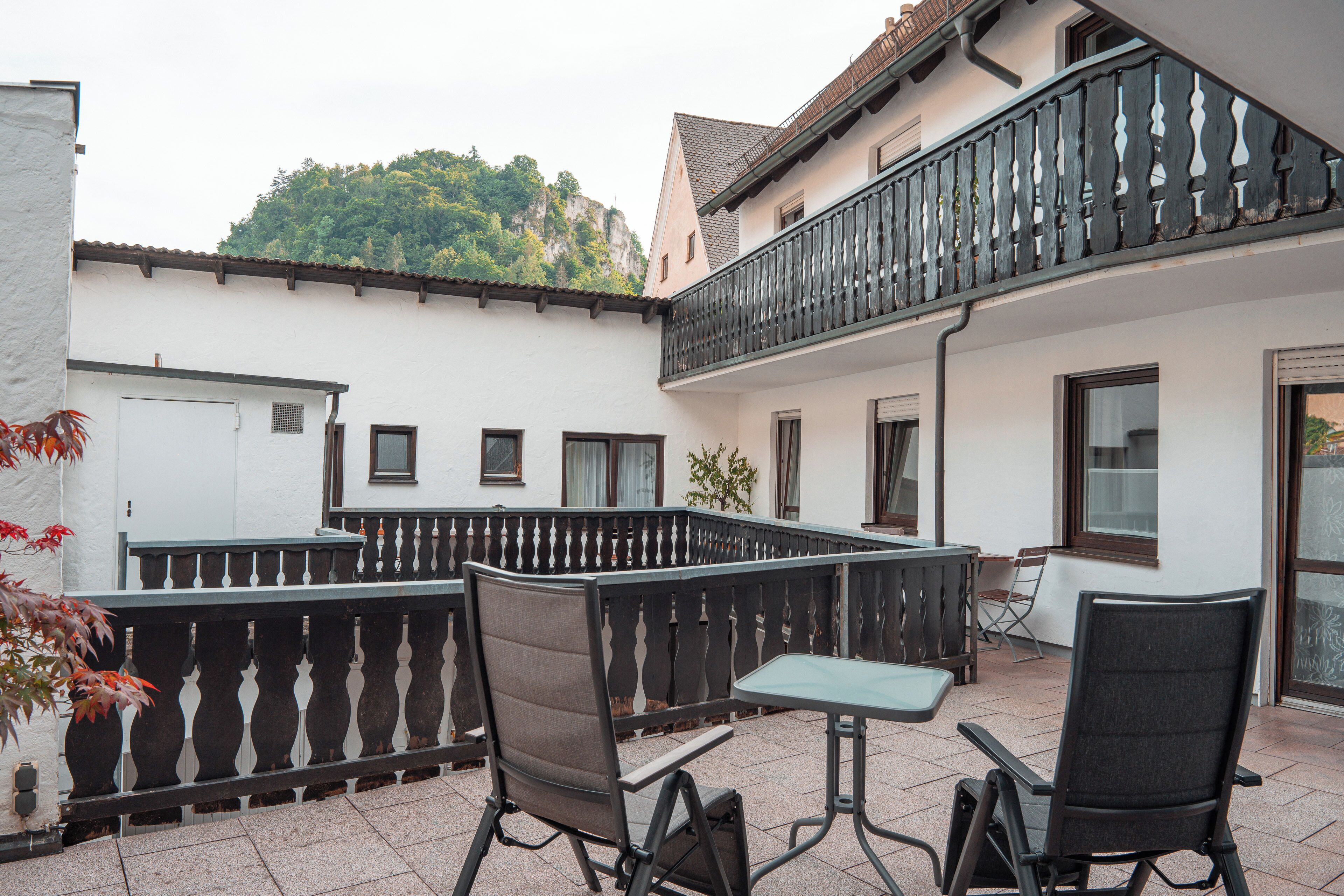 A courtyard with outdoor seating, a white building with a balcony, and a view of a forested hill.