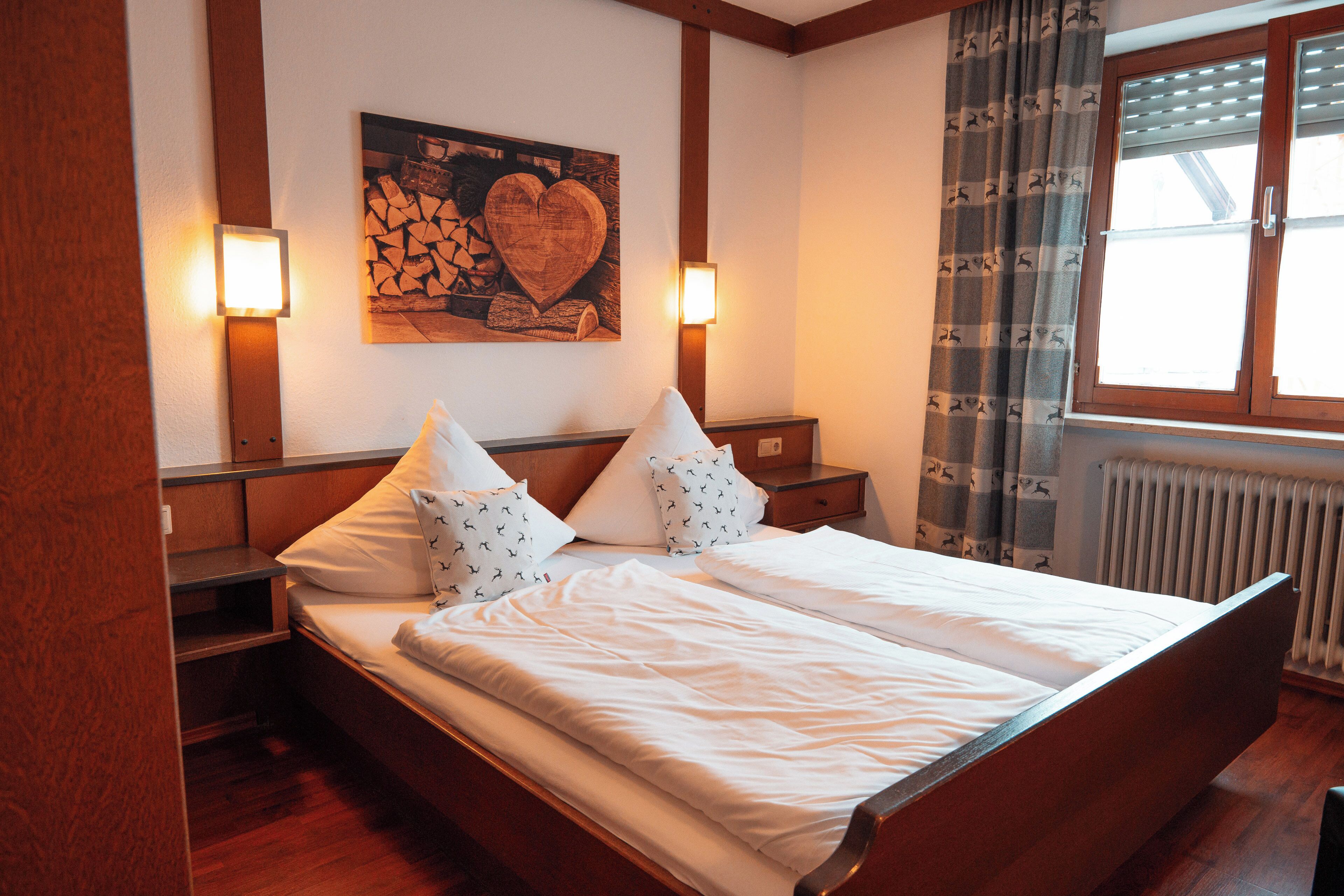 Una cama bien tendida con sábanas blancas, dos mesitas de noche con lámparas, un cabecero de madera y un cuadro de un corazón en la pared.