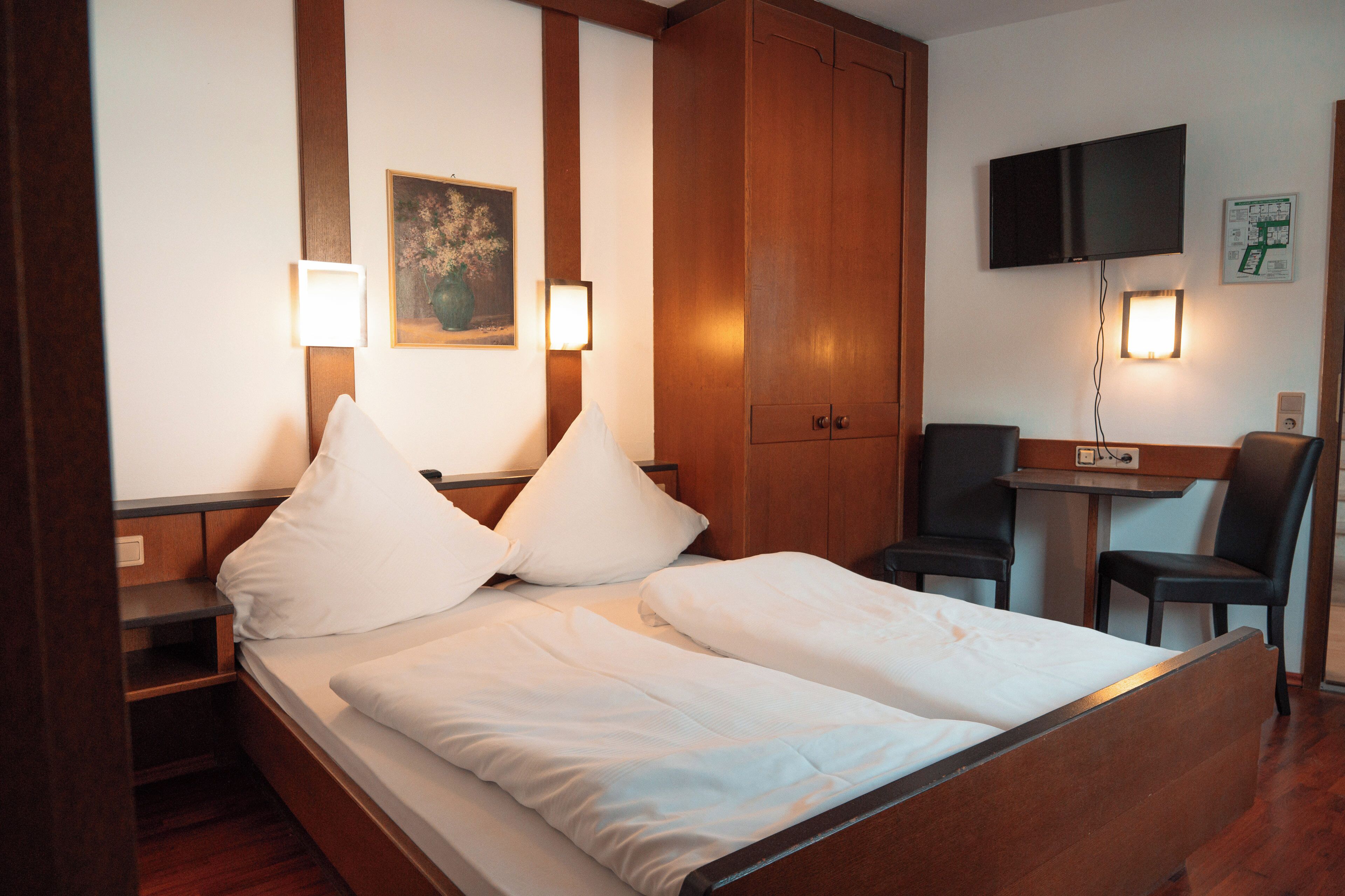 Une chambre d’hôtel avec un lit en bois, deux tables de chevet, un bureau avec une chaise, un téléviseur à écran plat fixé au mur et un tableau encadré au-dessus du lit.
