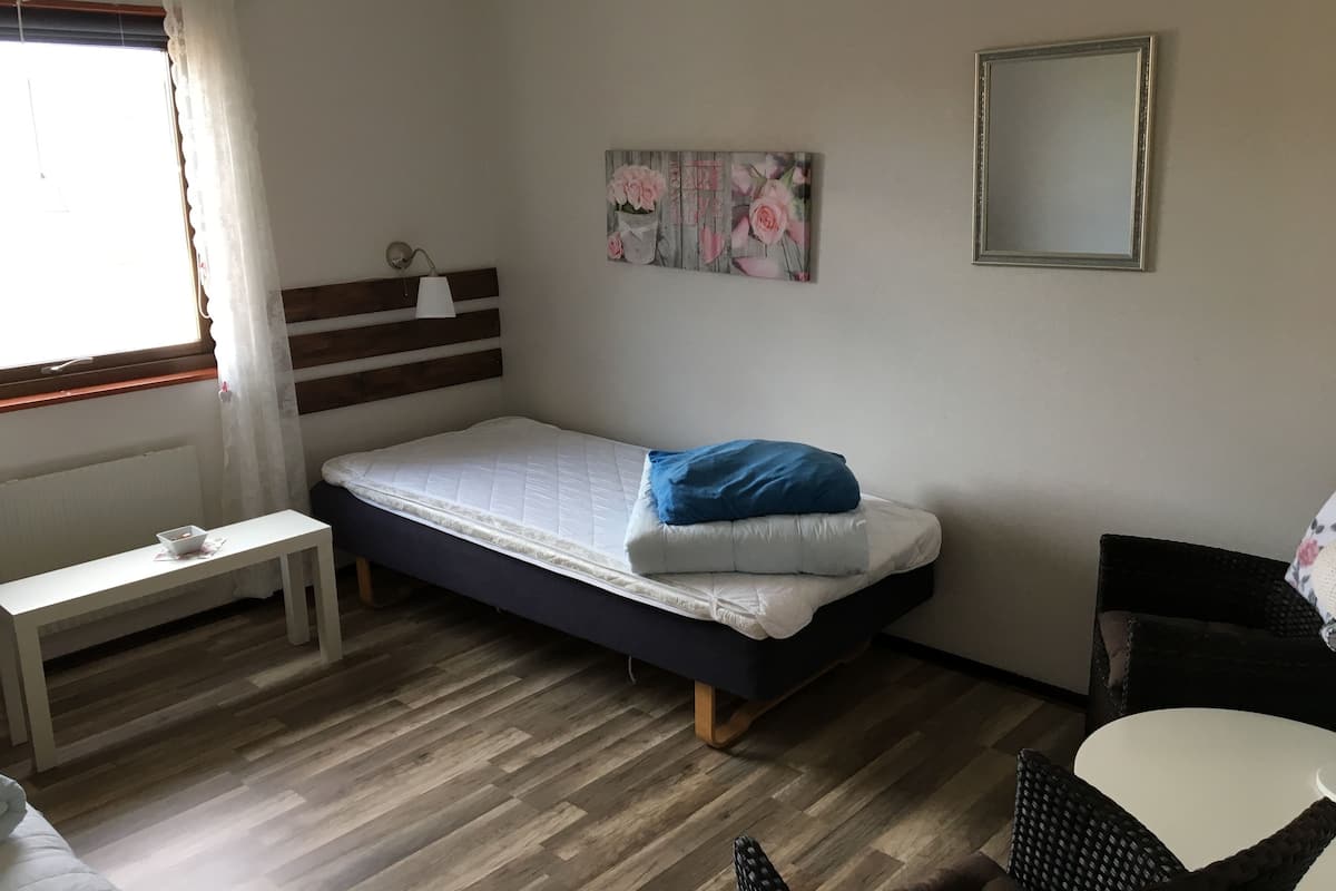 Une chambre avec un lit simple, une table de chevet, une chaise, une petite table, un miroir et une fenêtre avec des rideaux.
