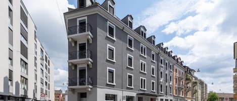 Un edificio de varios pisos con fachada moderna, una entrada visible en la planta baja y una vista de la calle con coches estacionados y cielo despejado.