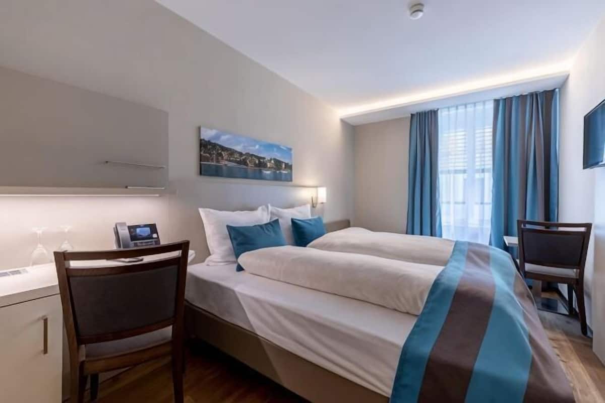 A hotel room with a bed, a desk, a chair, a TV, and a window with curtains.