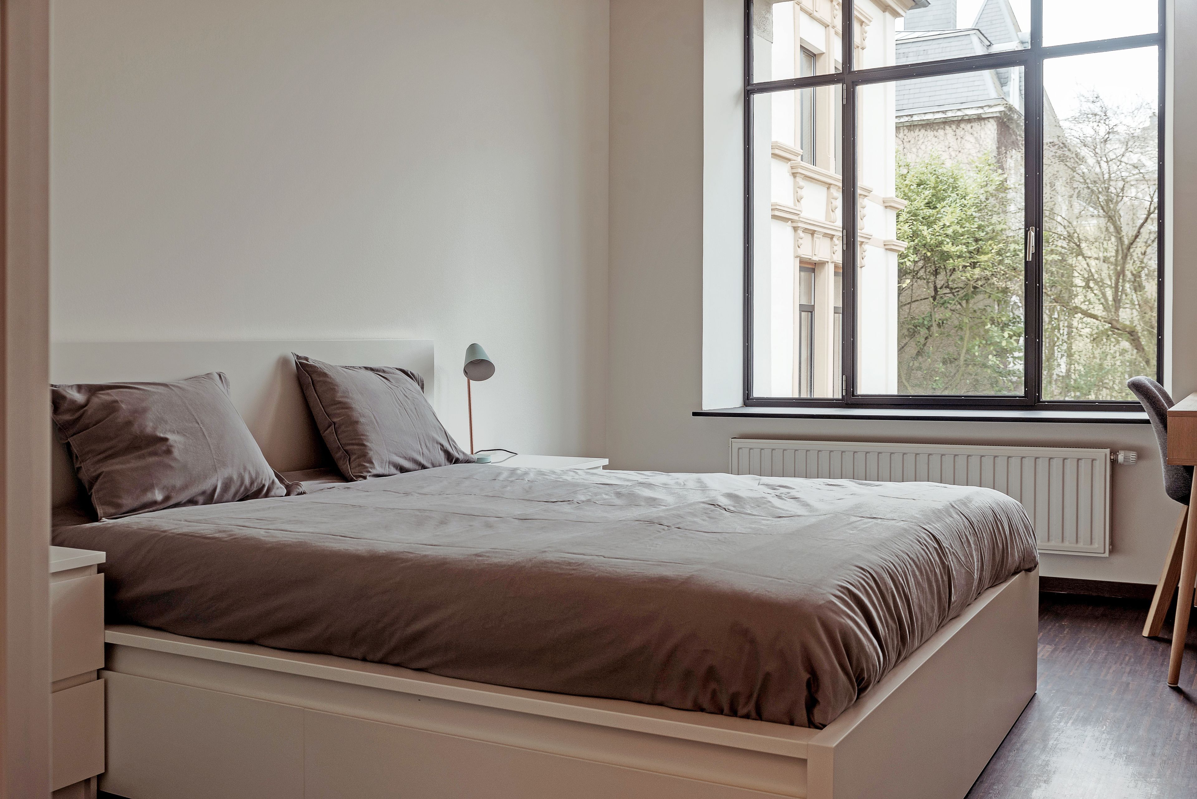 Un dormitorio con un ventanal grande, una cama, una mesita de noche y un radiador.
