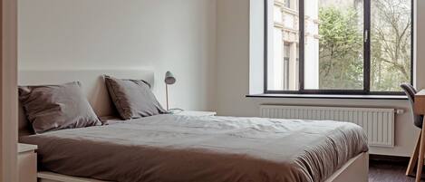 Un dormitorio con un ventanal grande, una cama, una mesita de noche y un radiador.