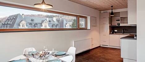 A modern kitchen with white cabinets, a dining area with a table set for four, and a large window offering a view of houses and trees.