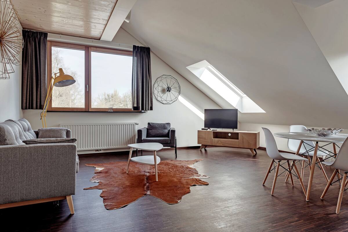 A modern loft apartment with a sloped ceiling, a large skylight, a cozy seating area with a sofa and armchairs, a dining table with chairs, and a flat-screen TV on a wooden cabinet.