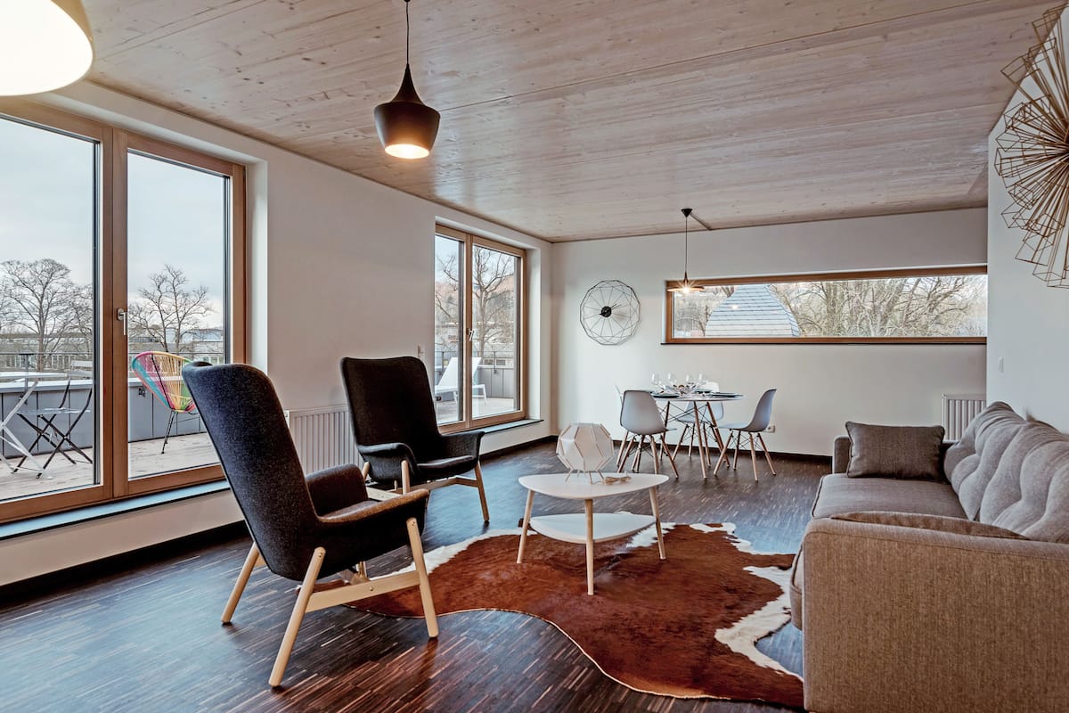 A modern living room with a wooden ceiling, large windows, a cowhide rug, a sofa, a chair, and a dining area with a table and chairs.