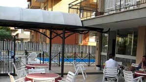 Outdoor seating with white chairs and red tablecloths, a covered area, and a pool in the background. - Hotel Mutacita (Rimini)
