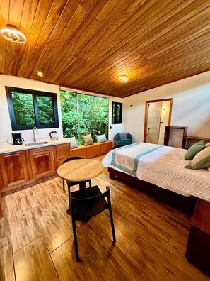 A bedroom with a wooden ceiling, a bed, a desk with a chair, a window, and a view of greenery. - Cabañas Hoja Verde (Monteverde)