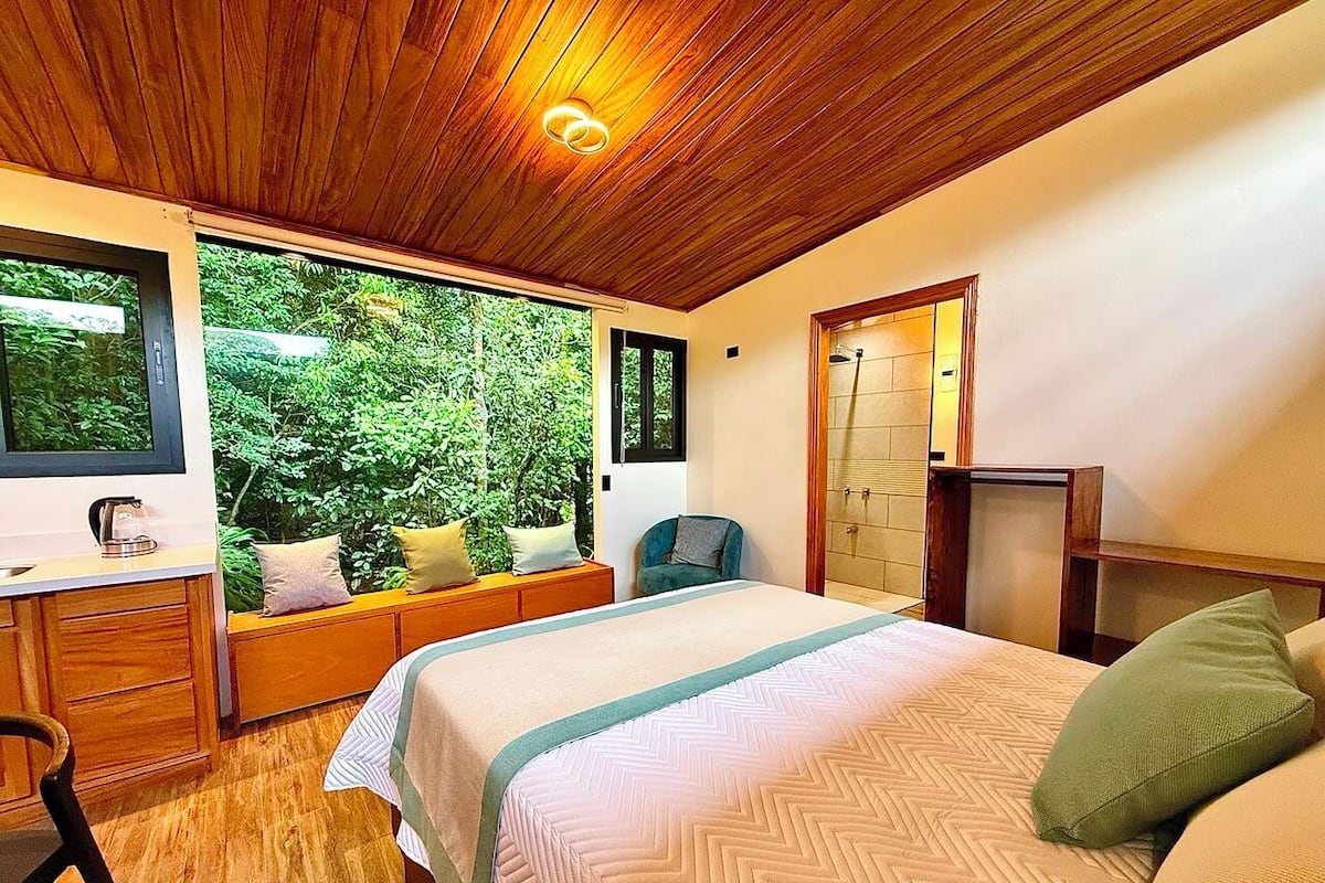 A bedroom with a bed, a desk, a chair, a window with a view of greenery, and a wooden ceiling.