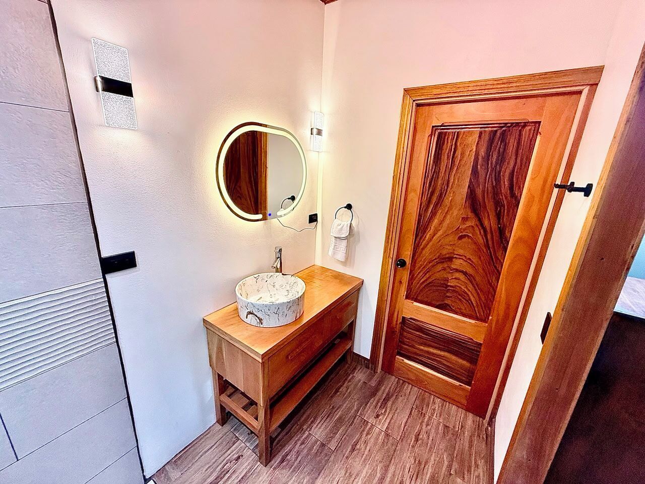 A bathroom with a wooden vanity, a round mirror with a light, and a wooden door.
