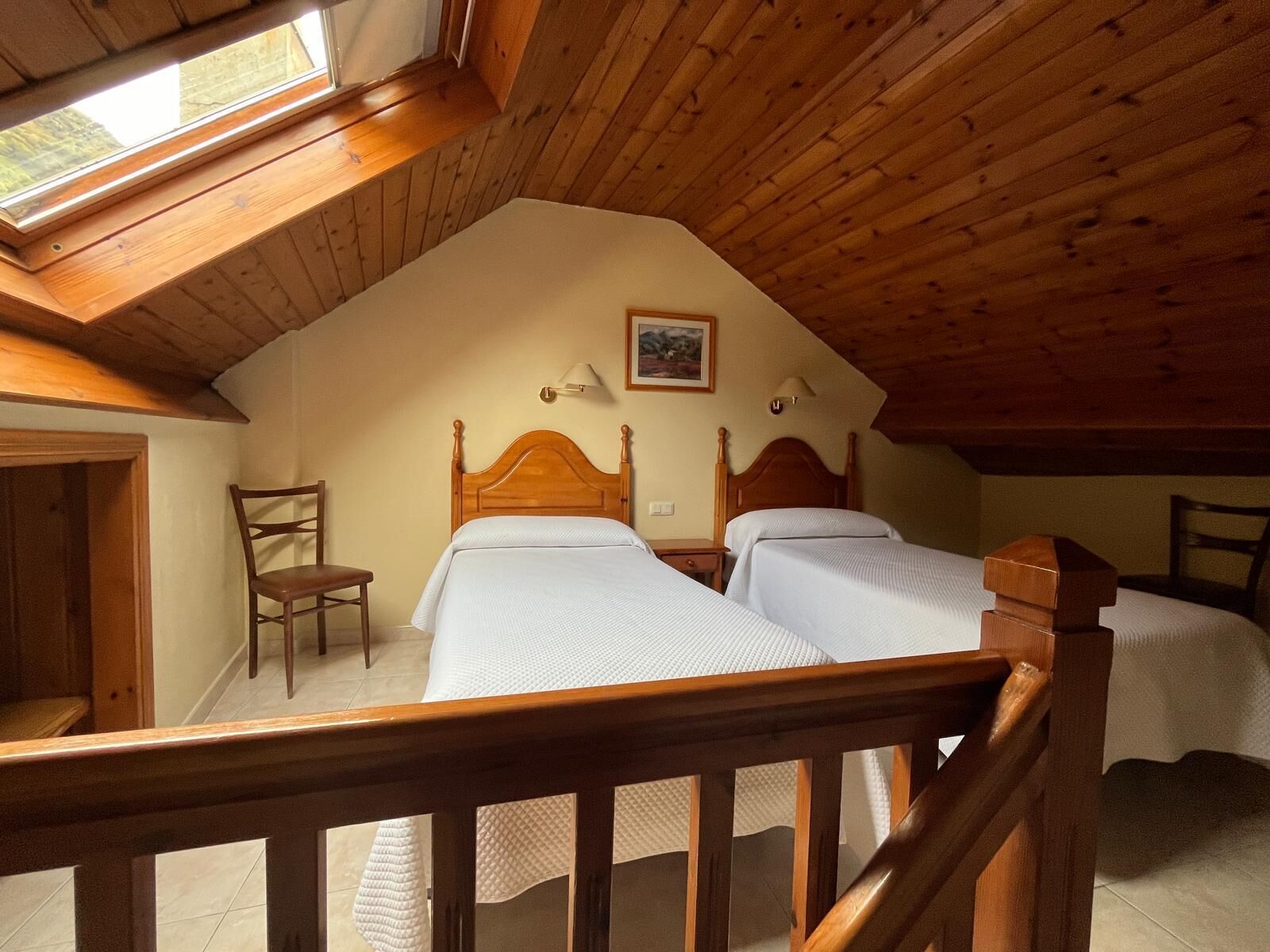 Un dormitorio con dos camas, cabeceros de madera, una silla de madera, una mesita y un cuadro en la pared.