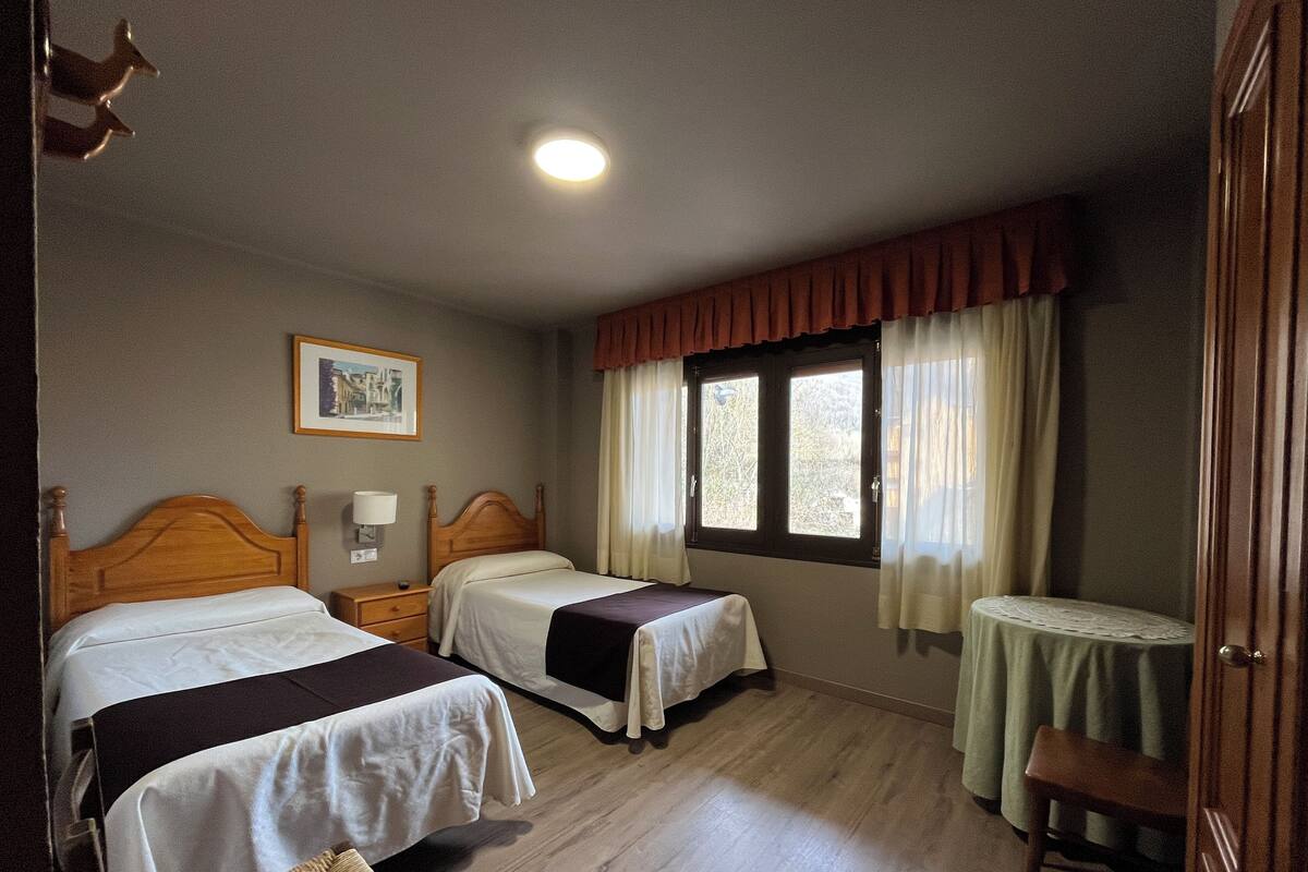 A hotel room with two beds, a small table, a chair, a window with curtains, a framed picture on the wall, and a ceiling light.
