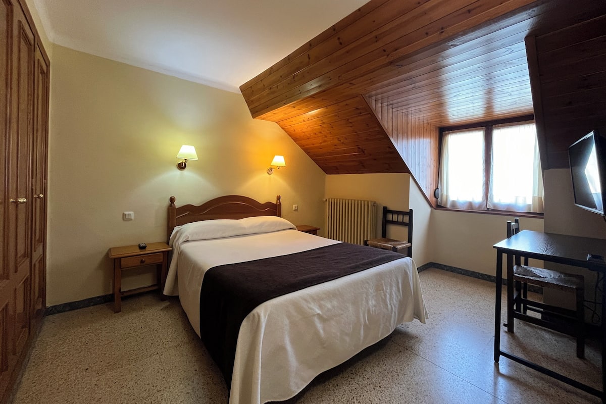 A bedroom with a bed, bedside tables, a chair, a desk, a television, and a window with blinds.