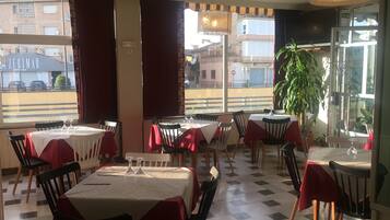 A restaurant interior with tables set for dining, a large potted plant, and a view of the street outside.