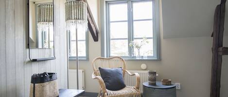 Une pièce comprenant un fauteuil en osier, un banc, une table ronde, un miroir et une fenêtre donnant sur des arbres.