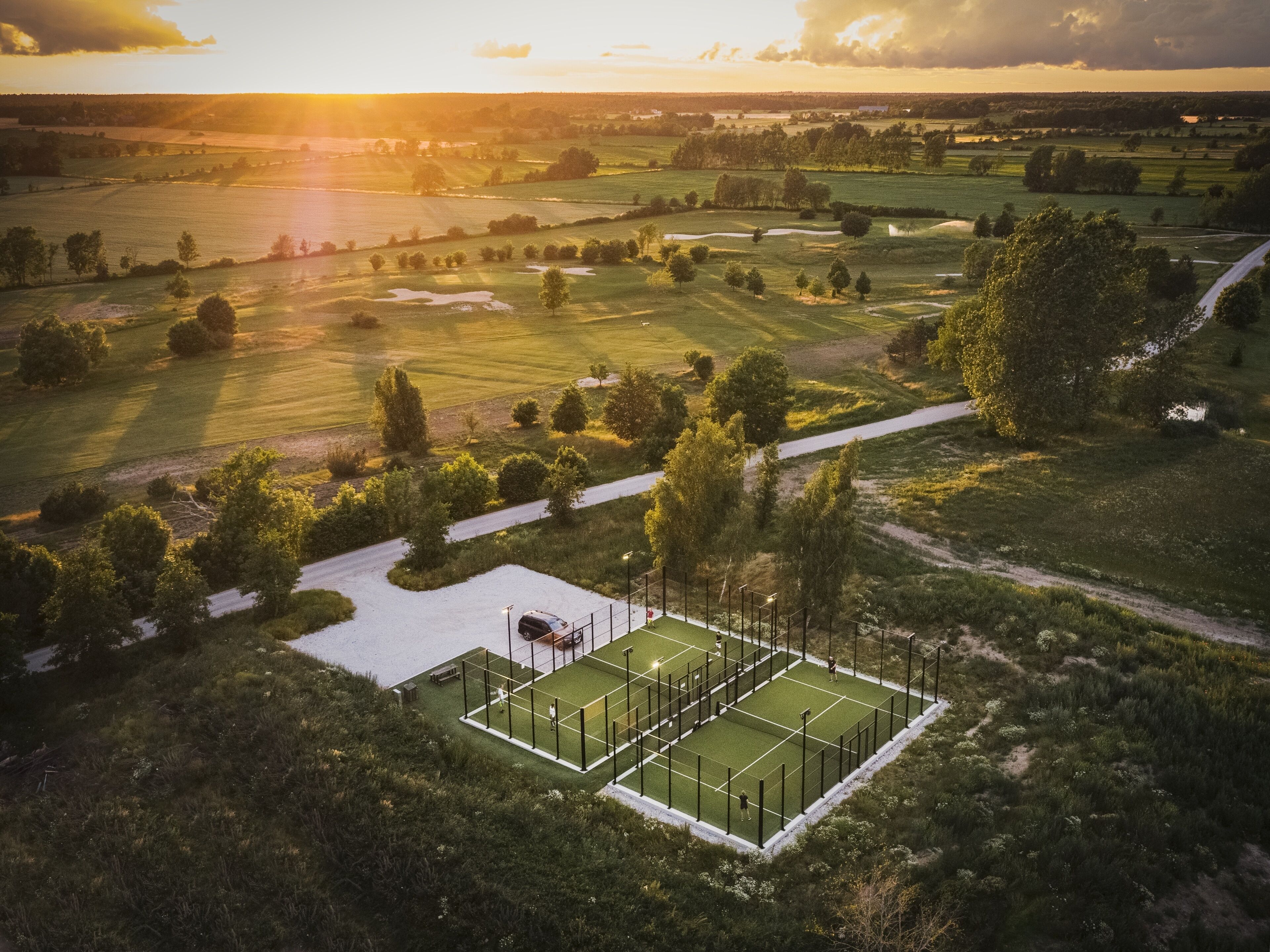 Une vue aérienne d’un court de tennis entouré d’une végétation luxuriante et de champs, avec un coucher de soleil en arrière-plan.
