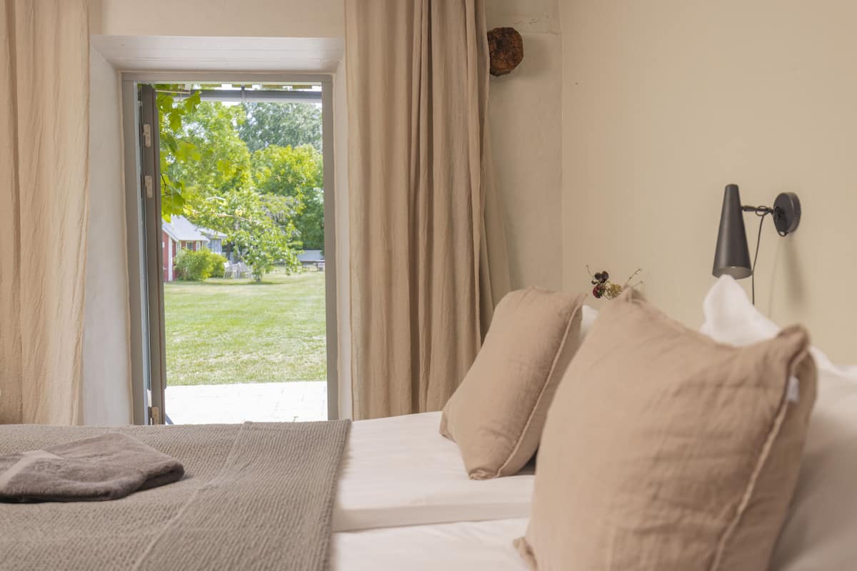 Quarto com cama, cortinas bege, vista para um jardim e luminária fixada na parede.