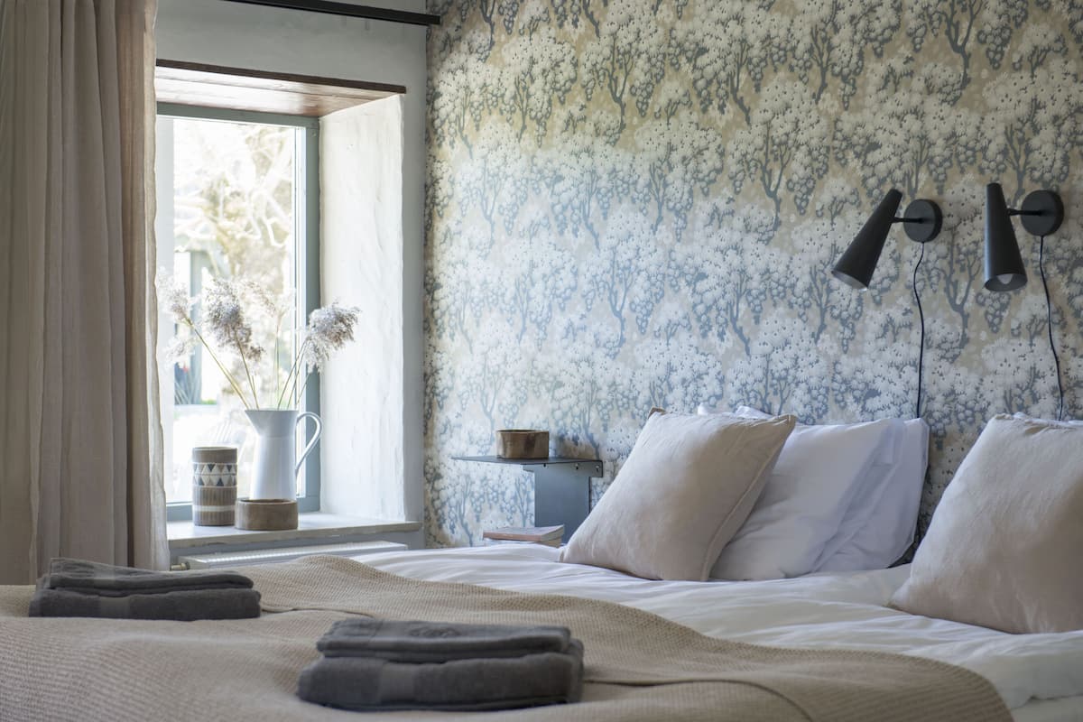 Um quarto com cama, almofadas, mesa de cabeceira com abajur, janela com cortinas e um vaso com flores.