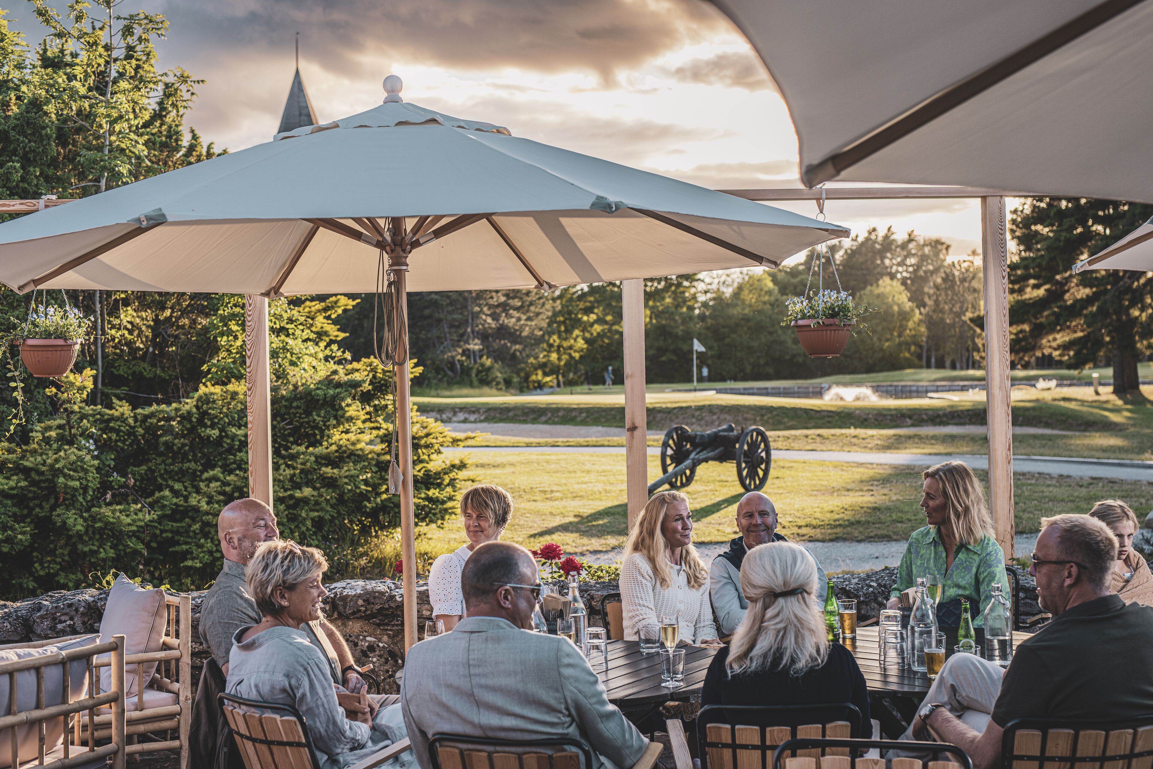 A group of people seated at an outdoor table under a large umbrella, with a golf cart and greenery in the background.