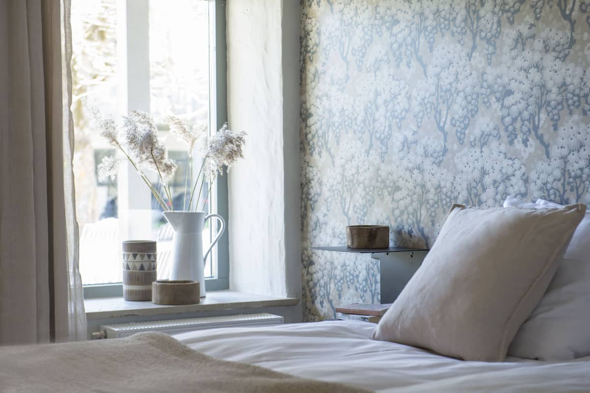 Quarto com cama, mesa de cabeceira, janela com cortinas e um vaso com flores secas.