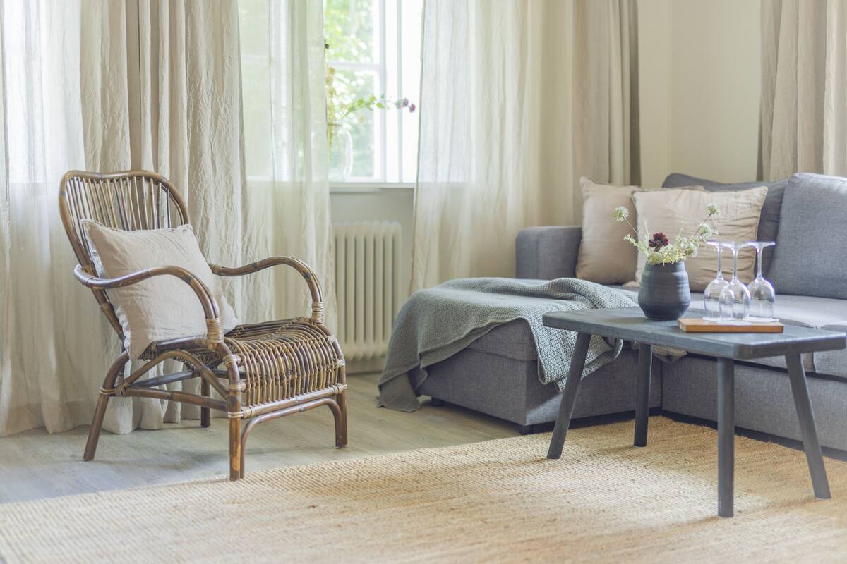 Quarto com uma cadeira de vime, um sofá cinzento e uma mesa de centro com um vaso e copos.