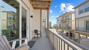 A wooden deck with a chair and a doormat, overlooking a street with buildings and a parked car. - Little Lagoon Cottages (Gulf Shores)