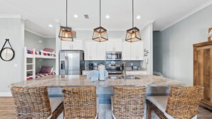A modern kitchen with a central island, white cabinets, stainless steel appliances, and wicker chairs. - Little Lagoon Cottages (Gulf Shores)