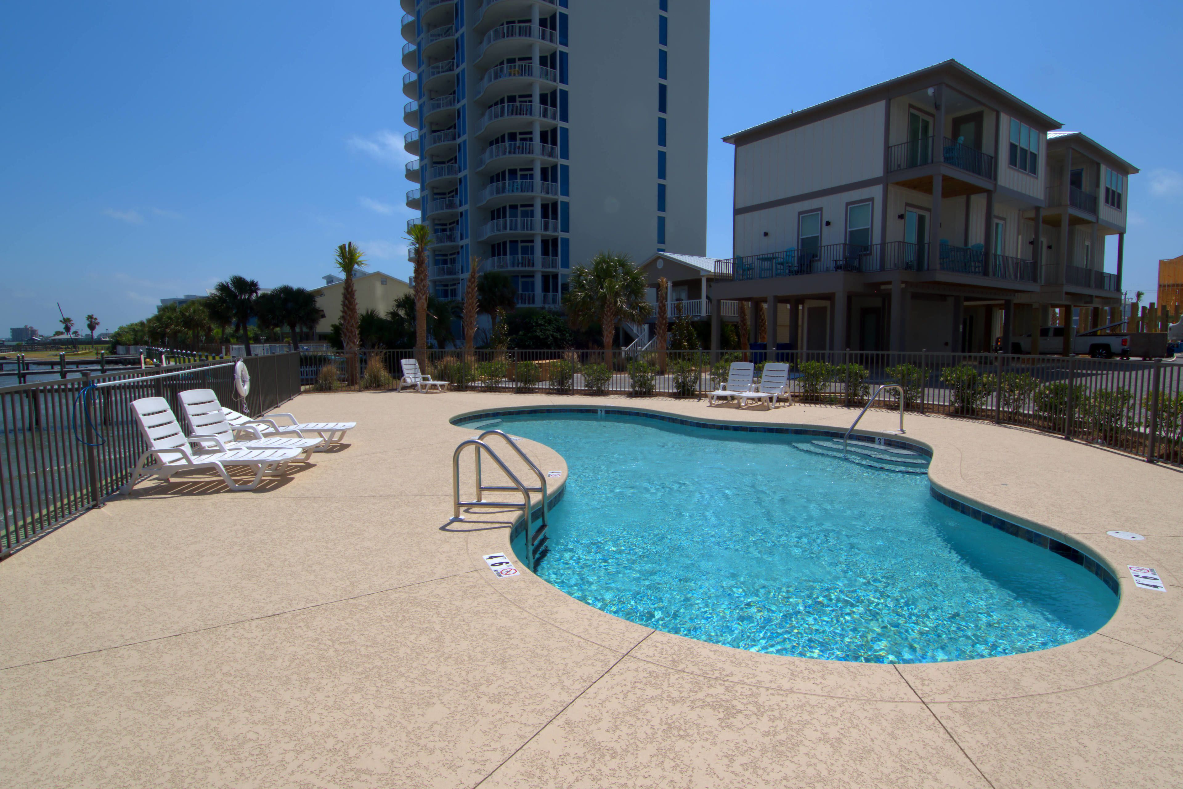 Una piscina con sillas de descanso, un puesto de socorrista y un edificio alto al fondo.