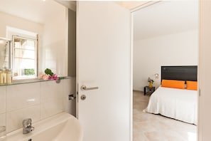 A hotel room with a bathroom, a bed with orange and black bedding, and a small bedside table with a vase.