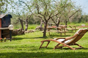 Un fauteuil de jardin en bois, placé dans un espace extérieur verdoyant, avec des tables de pique-nique et un barbecue en arrière-plan.