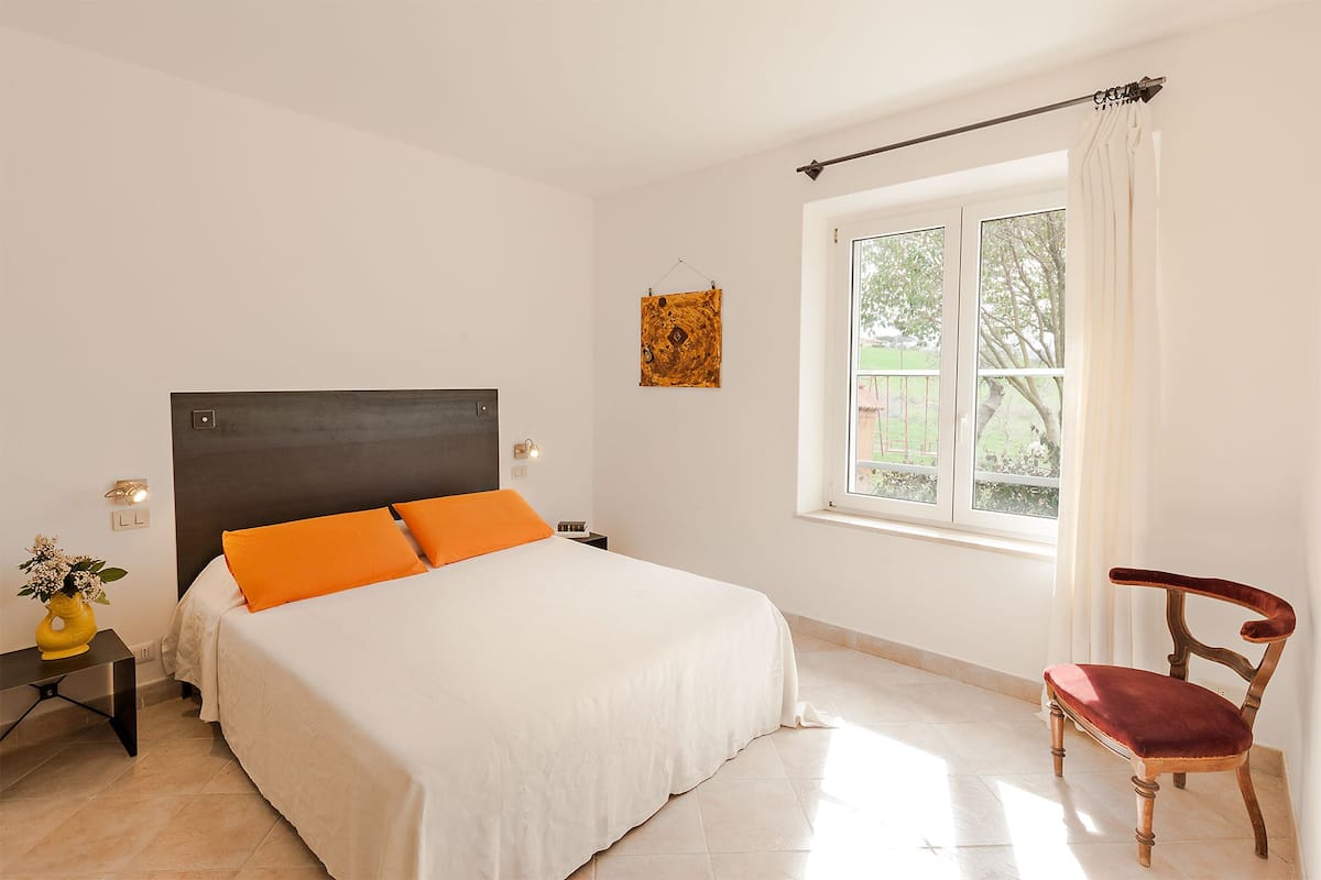 Ein Schlafzimmer mit einem Bett, einem Nachttisch, einem Sessel, einem Fenster mit Vorhängen und einem Wandbild.