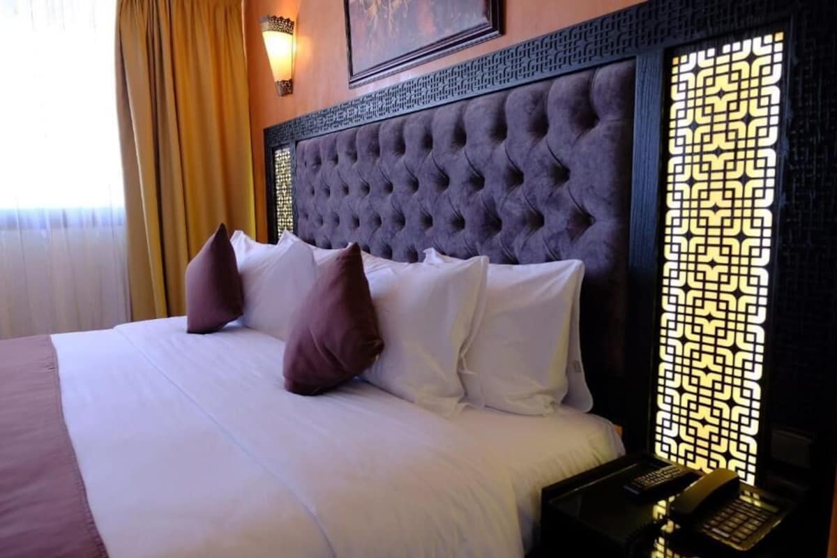 Una habitación de hotel con un cabecero capitonado, una cama con ropa blanca y dos almohadas, un teléfono en la mesita de noche y un panel decorativo en la pared.