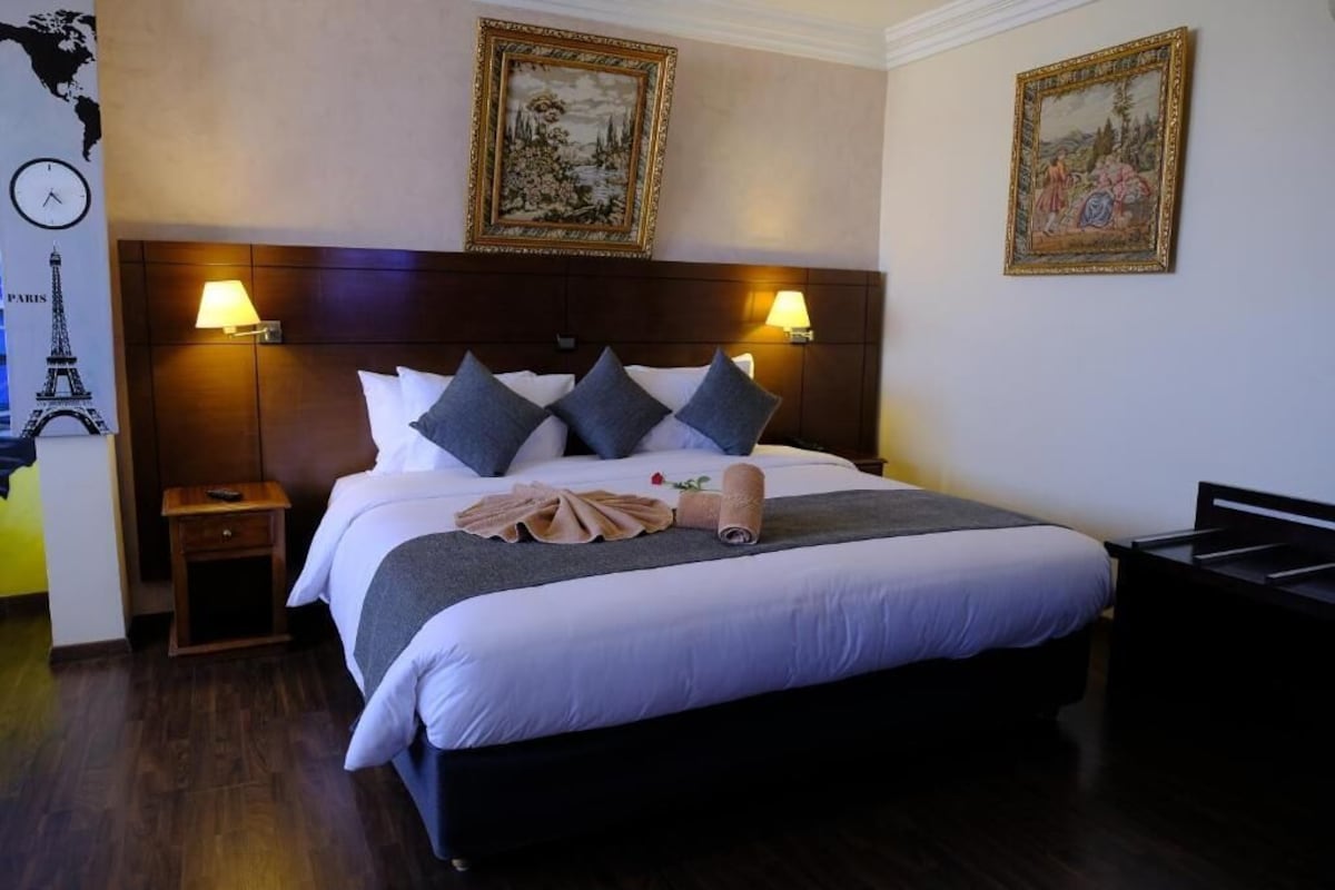 Una habitación de hotel con una cama grande, dos lámparas de noche, una mesita de noche, un cabecero de madera y dos cuadros en la pared.
