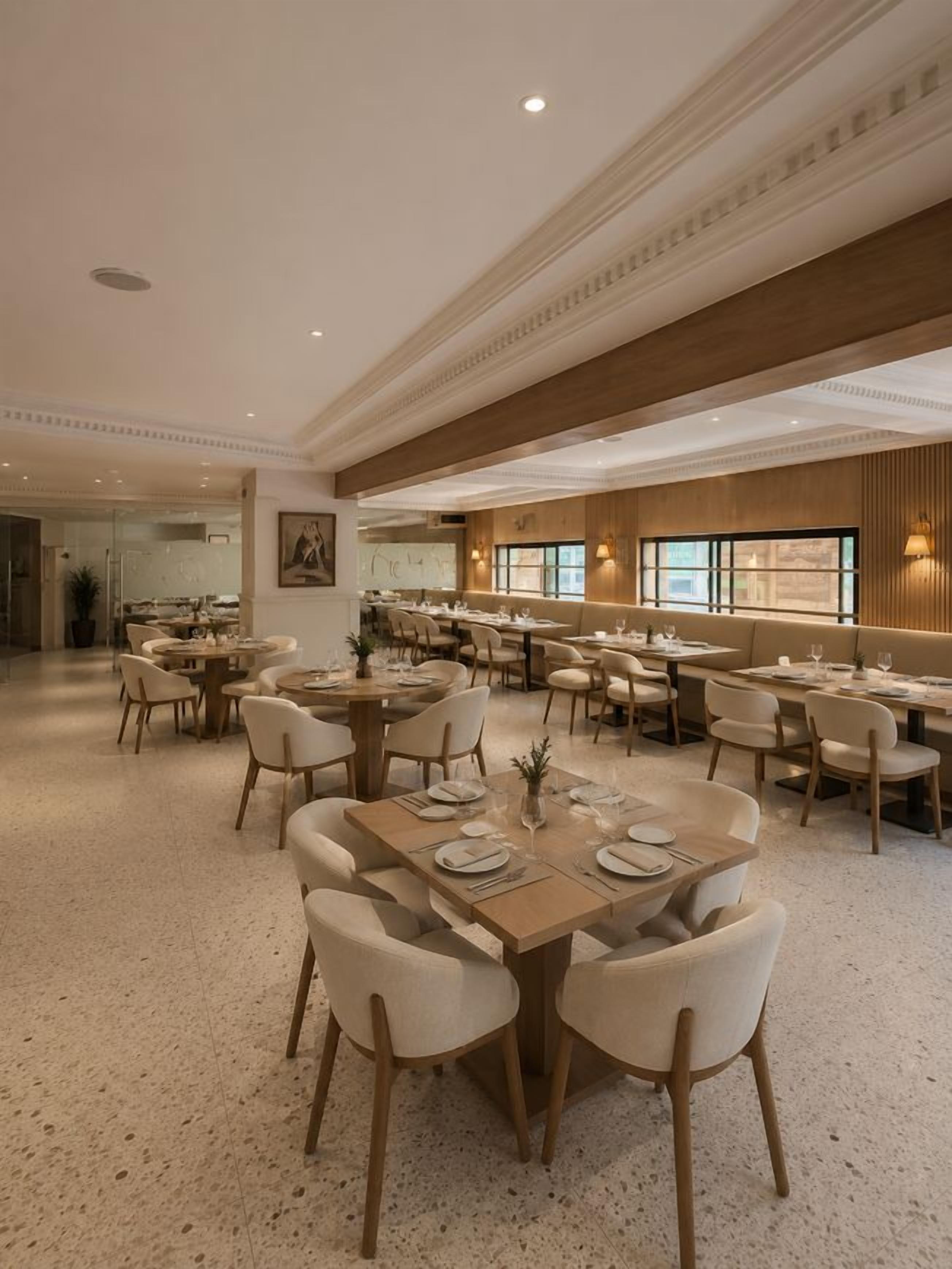 Un vaste espace dédié à la salle à manger, avec de nombreuses tables et chaises, un plafond haut orné de moulures décoratives et de grandes fenêtres laissant entrer la lumière naturelle.